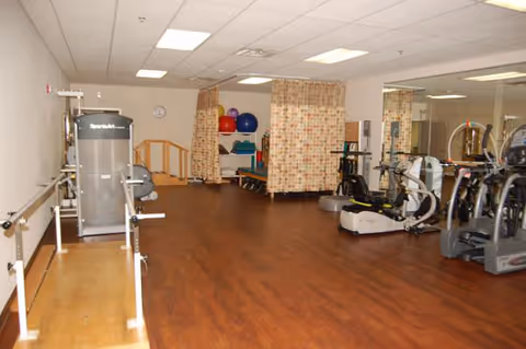 Interior view of a rehabilitation gym with exercise equipment including stationary bikes, parallel bars, therapy balls, and treatment tables separated by curtains. The room has wood flooring and fluorescent ceiling lights.