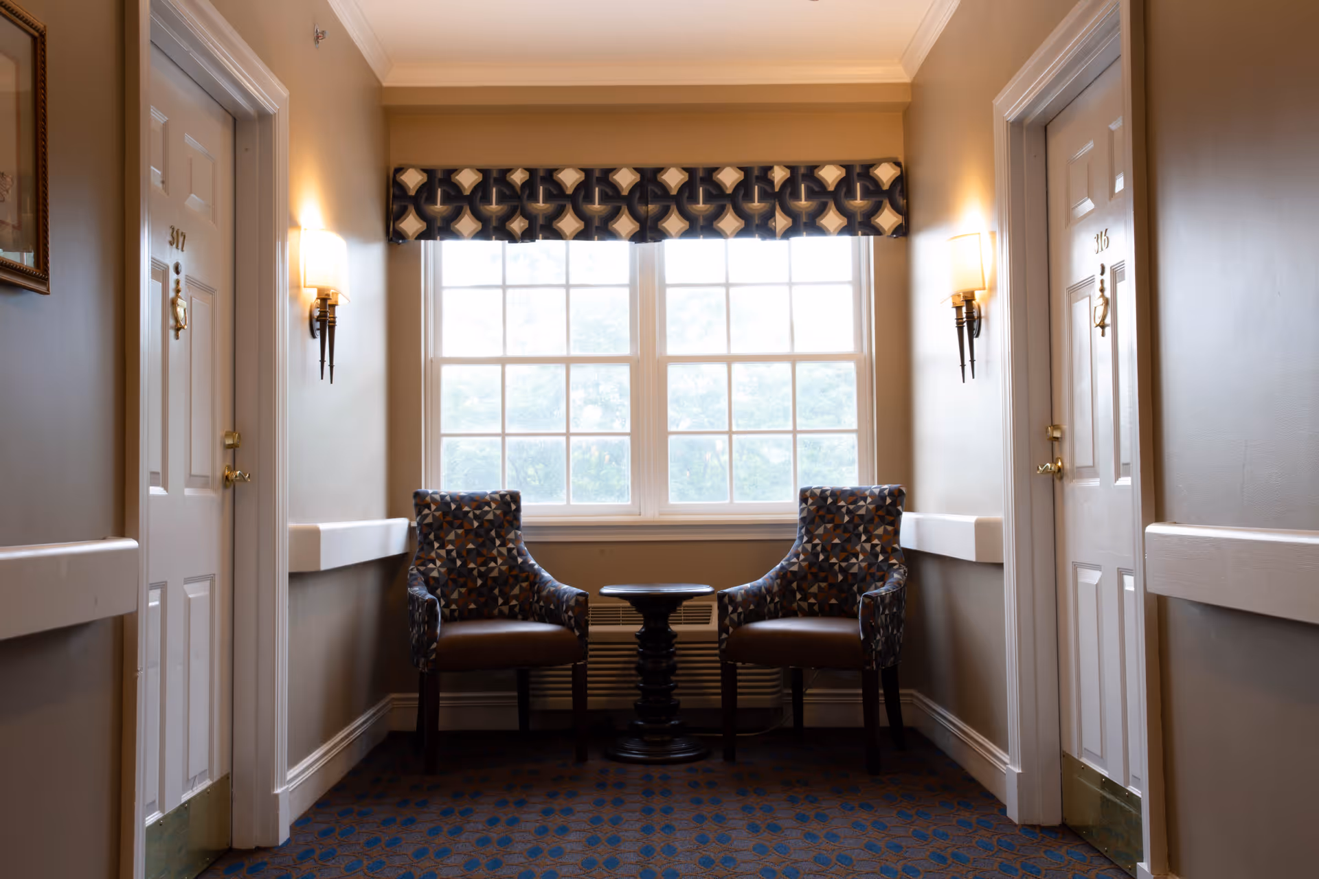 A small sitting area in a hallway with two patterned armchairs and a round black table between them, positioned in front of a large window with a decorative valance. The hallway has beige walls, two white doors labeled 316 and 317, wall-mounted light fixtures, and a carpeted floor with a blue and brown pattern.