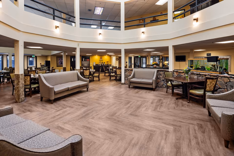 Spacious senior living community common area with multiple seating arrangements including sofas and chairs around tables. The room features a high ceiling with a second-floor balcony railing, wood-patterned flooring, and warm lighting. There is a fish tank on the right side and a coffee station in the background.