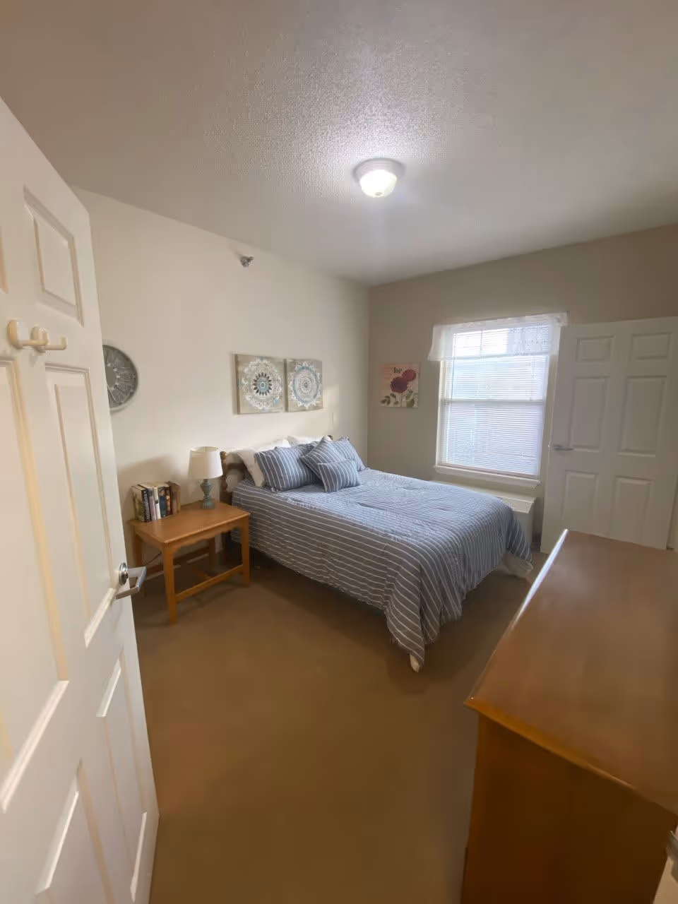 A bedroom with a neatly made bed featuring blue and white striped bedding and matching pillows. There is a wooden nightstand with a lamp and some books on it next to the bed. The room has beige walls with two decorative wall hangings above the bed and a floral picture near a window with blinds and a white valance. A wooden dresser is visible in the foreground, and the door to the room is open.