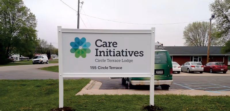 Outdoor view of a white signboard with the logo and text 'Care Initiatives Circle Terrace Lodge 155 Circle Terrace' placed on a grassy area near a parking lot with several parked cars and a building in the background.