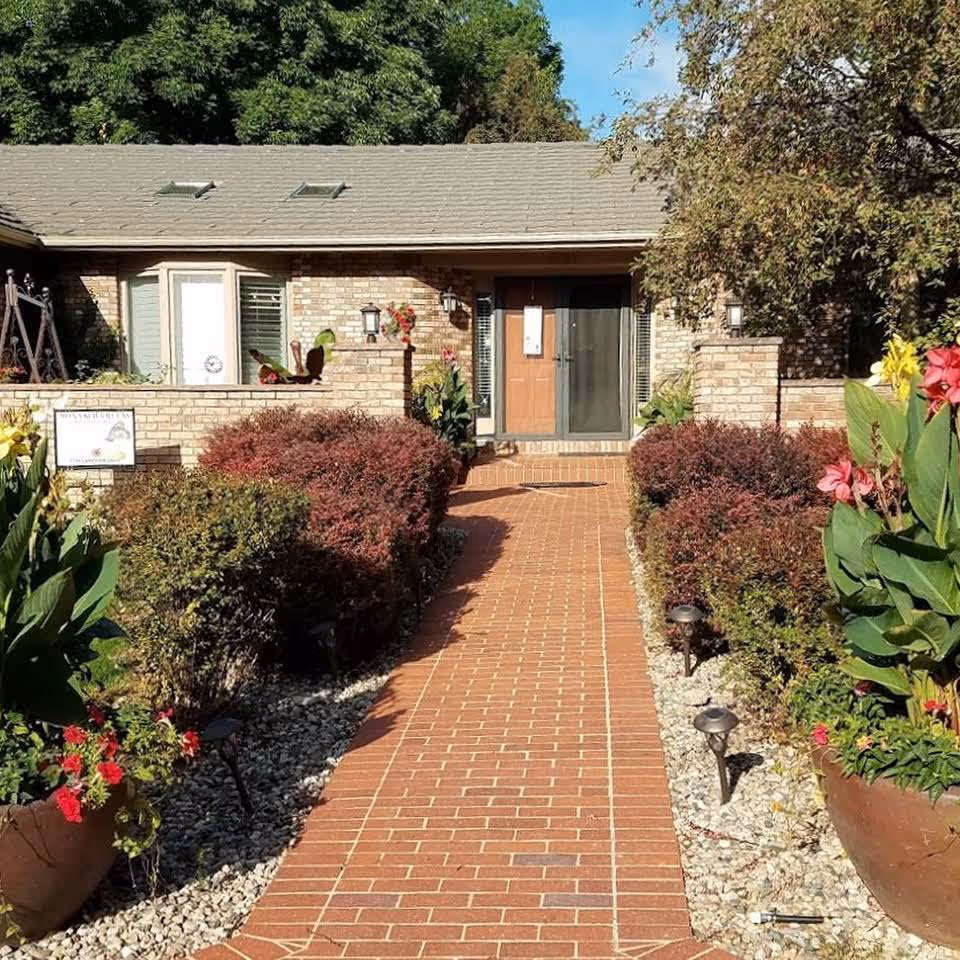 A brick walkway leading to the entrance of a single-story brick building surrounded by well-maintained bushes and large flower pots with colorful flowers. The building has a brown door with a glass storm door and small windows on either side. Trees and greenery are visible in the background under a clear blue sky.