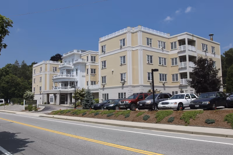 Exterior view of a multi-story beige and white building with balconies and a parking lot in front. The building is surrounded by greenery and trees under a clear blue sky. A sign near the entrance reads 'Riverview Rehabilitation & Healthcare Center.'