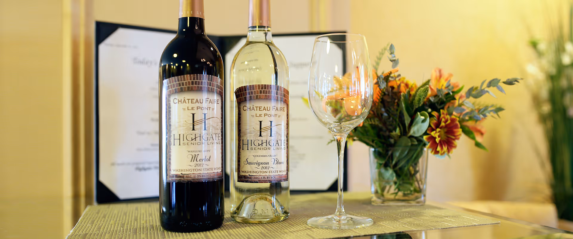 Two bottles of wine, one red and one white, labeled Highgate Senior Living, placed on a table next to an empty wine glass and a small vase with colorful flowers. A framed menu or document is visible in the background.