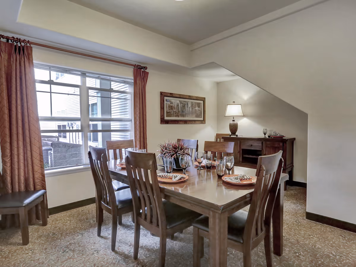 Interior dining room with a wooden table set for six, matching chairs, a sideboard with a lamp, and a large window with curtains.