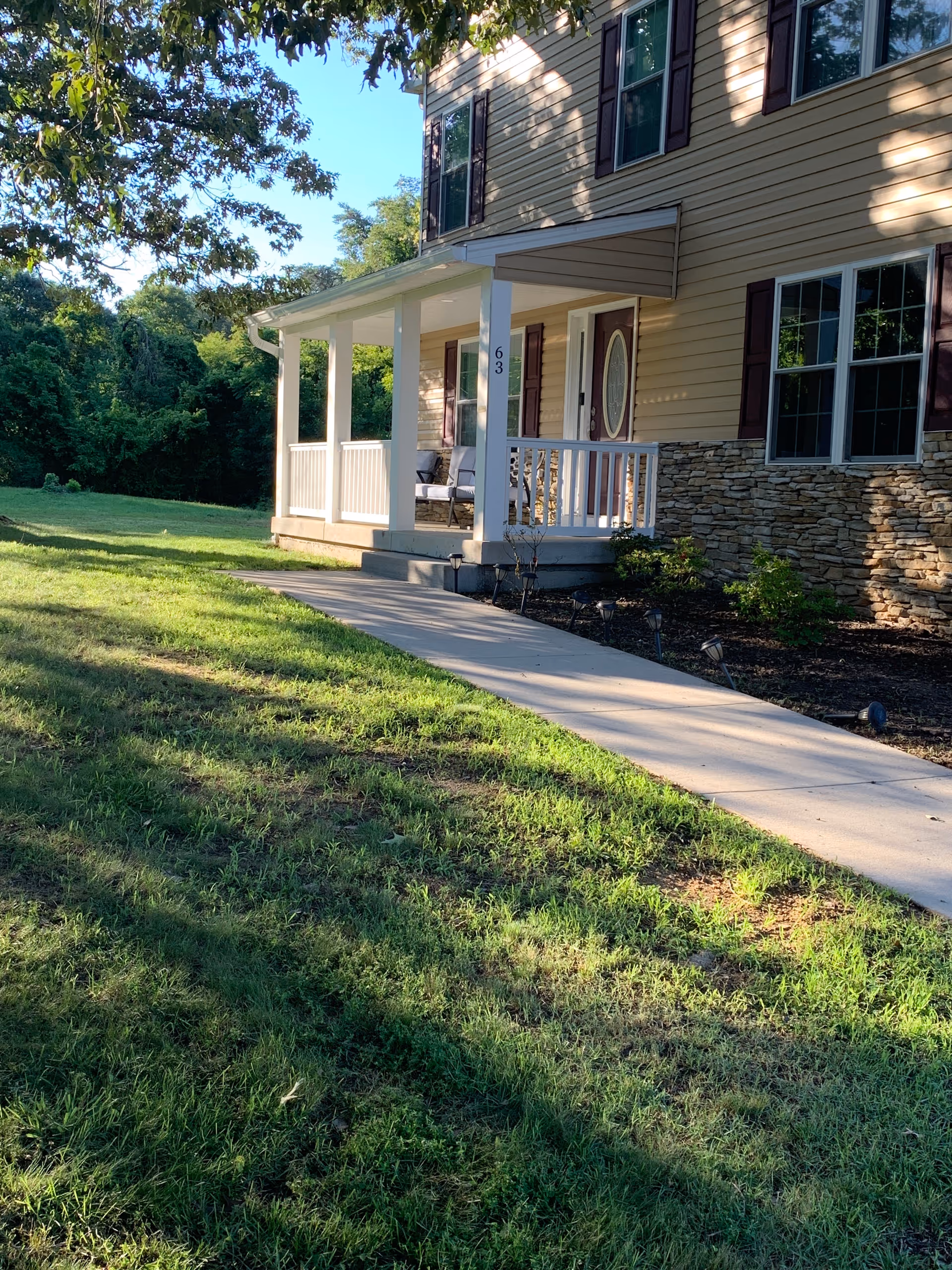 Front porch of a two-story beige house with a paved walkway, white columns, stone siding and a grassy lawn.