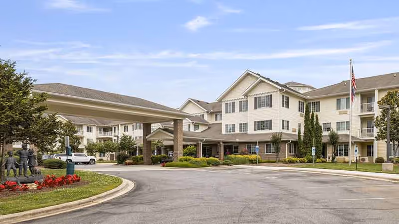 Exterior view of Oak Park Retirement facility showing a large multi-story building with beige siding and multiple windows. There is a covered entrance with pillars, a driveway, landscaped greenery, and an American flag on a flagpole. A statue of children playing is visible on the left side near the entrance.