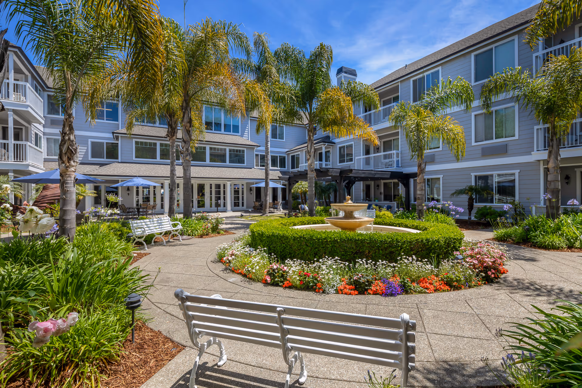 Outdoor courtyard area of a senior living facility with a central fountain surrounded by colorful flowers and neatly trimmed bushes. There are palm trees, white benches, and patio tables with blue umbrellas. The building surrounding the courtyard is three stories tall with balconies and large windows under a clear blue sky.