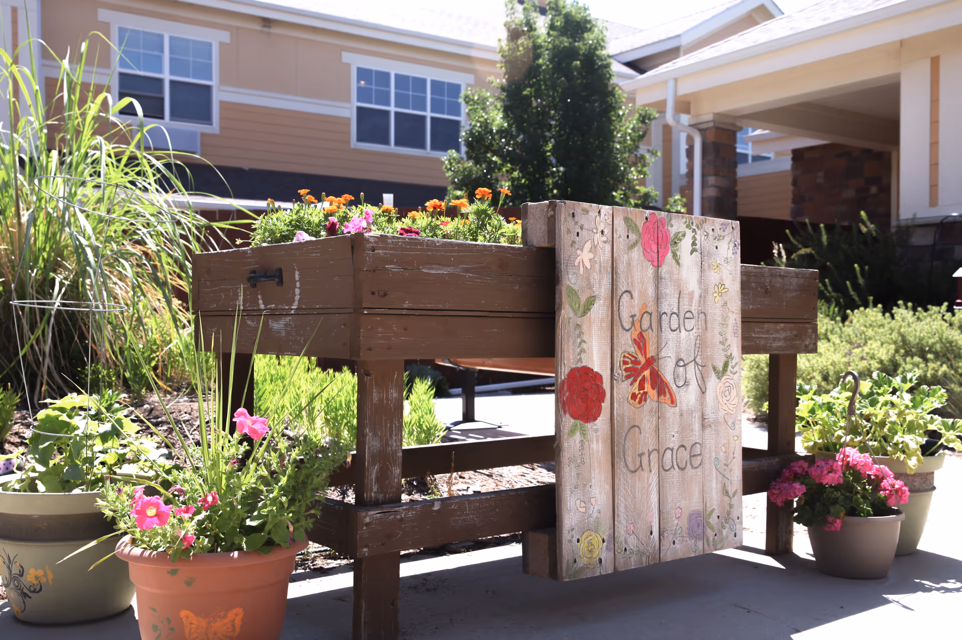 A raised wooden garden bed filled with colorful flowers and surrounded by potted plants. A wooden sign attached to the garden bed reads 'Garden of Grace' with painted flowers and a butterfly. In the background, there is a building with beige siding and white-trimmed windows under a clear sky.