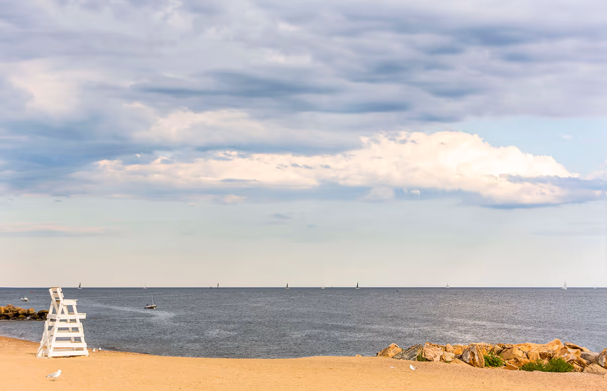 A sandy beach with a white lifeguard chair on the left side, calm ocean water with several small sailboats in the distance, and a partly cloudy sky overhead. There are rocks and some greenery on the right side of the beach.