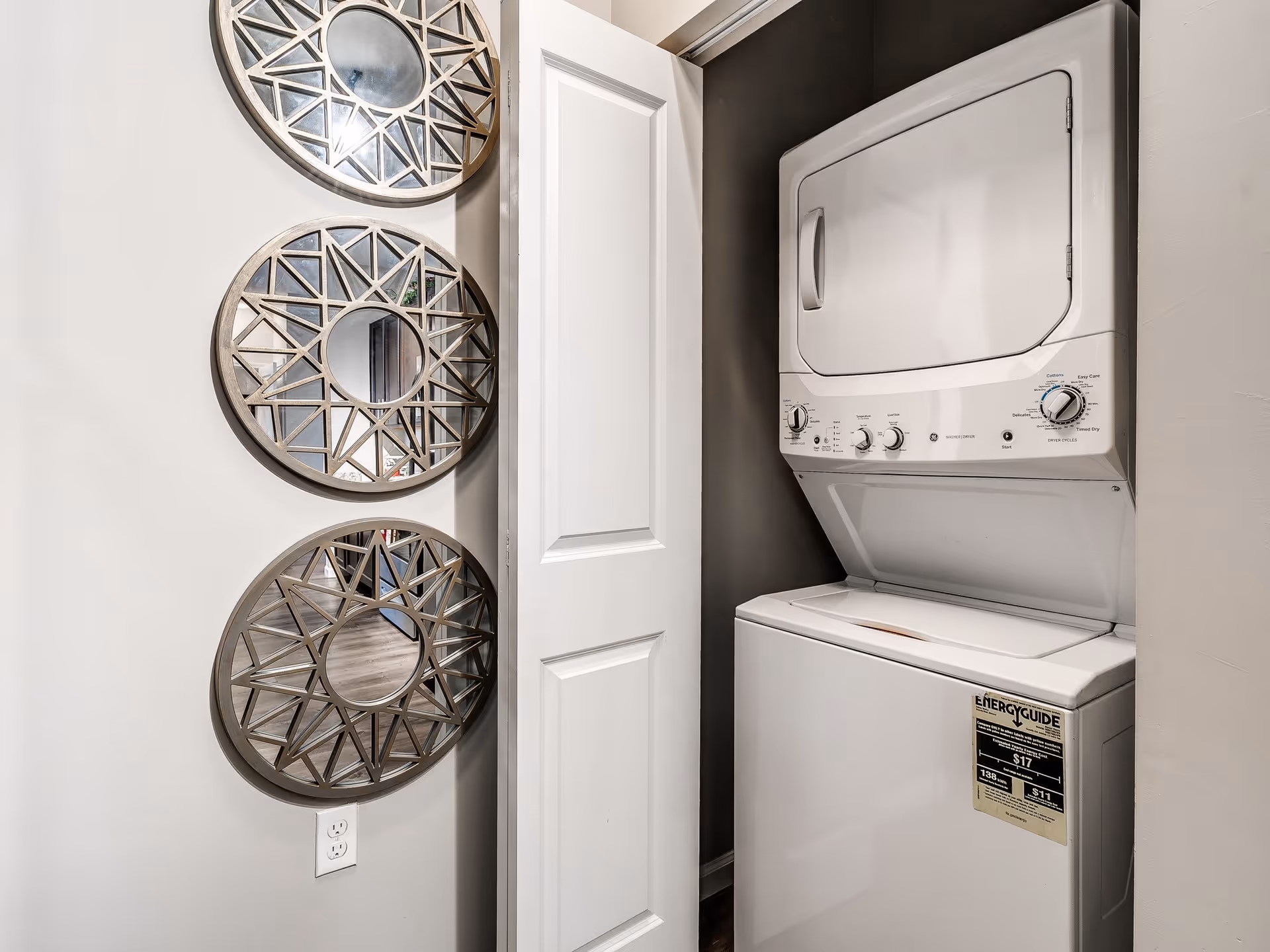 A small laundry closet with a stacked white washer and dryer unit. The closet has a white bi-fold door partially open. On the adjacent wall, there are three decorative round mirrors with geometric patterns arranged vertically. A power outlet is visible below the mirrors.