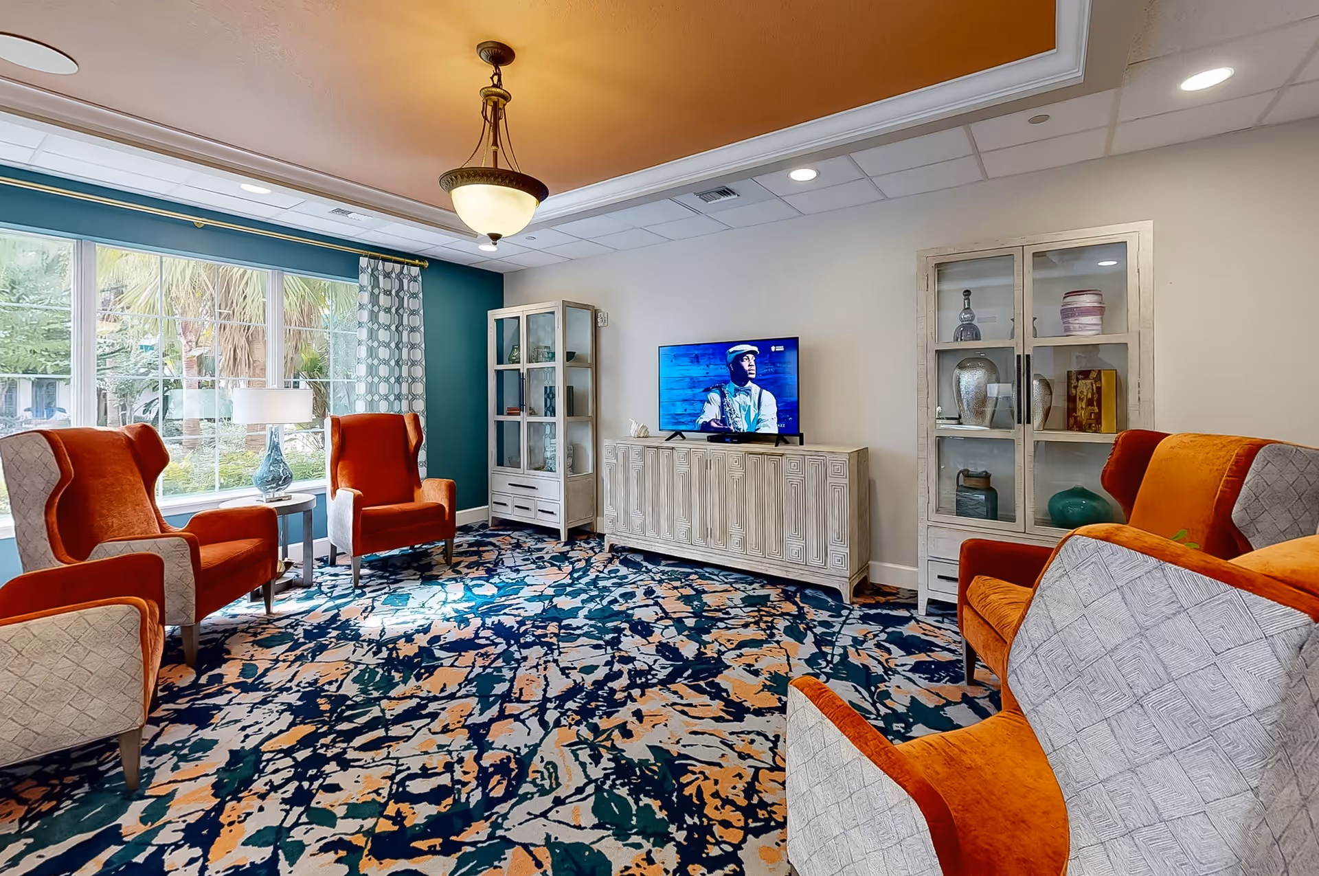 A bright living room with four orange and beige armchairs arranged around a patterned carpet. There is a large window with curtains letting in natural light, a white cabinet with a TV on top, and two glass display cabinets with decorative items inside. The ceiling has recessed lighting and a central hanging light fixture.