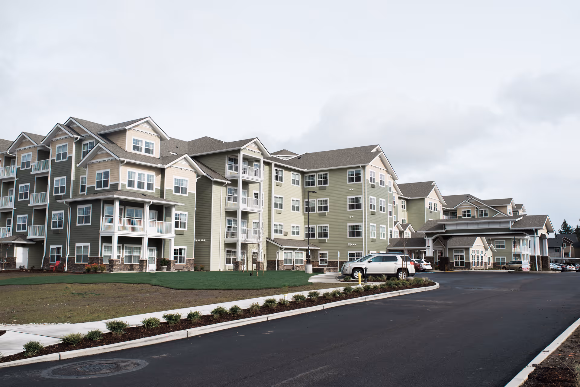 Exterior view of a large, multi-story senior living facility with green and beige siding, multiple balconies, and a covered entrance. Several cars are parked near the entrance along a paved driveway, with landscaped bushes and a grassy area in front.