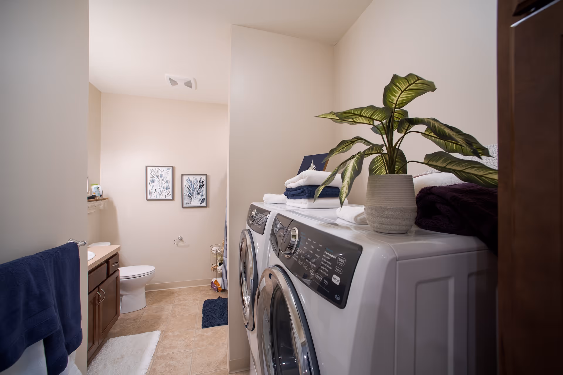 A bathroom with a toilet, sink with wooden cabinets, and a washing machine and dryer side by side. Towels and a potted plant are placed on top of the machines. Two framed botanical prints hang on the wall above the toilet. The floor is tiled and there are bath mats on the floor.