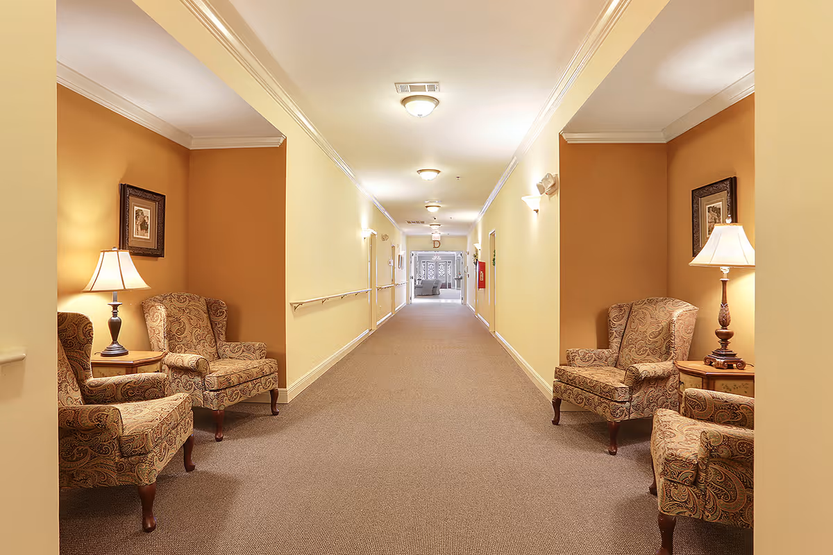 Well-lit interior hallway of a senior living facility with seating alcoves containing patterned armchairs and lamps.