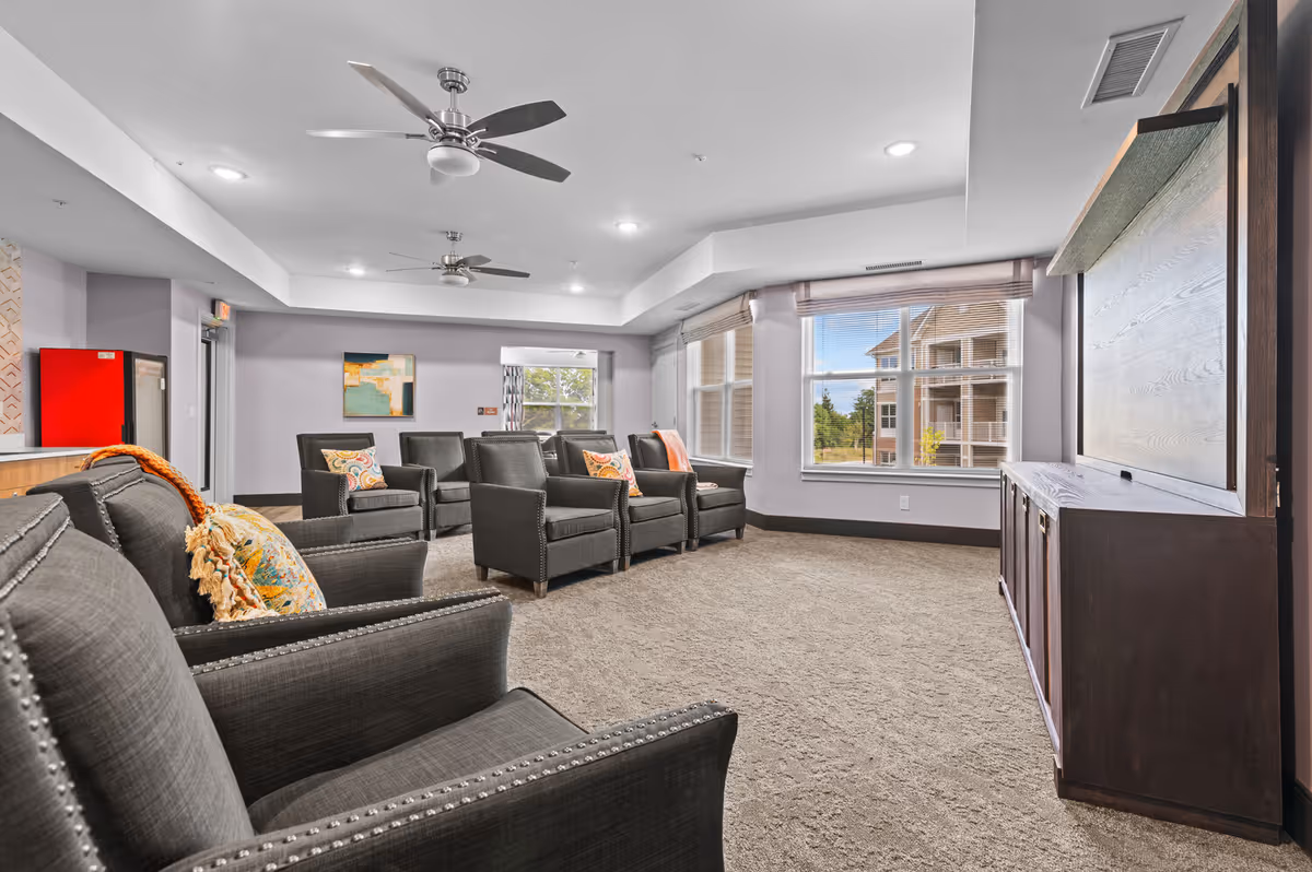 A spacious common living area with multiple gray armchairs arranged in rows facing a large wooden cabinet with a closed panel, large windows letting in natural light, ceiling fans, and a carpeted floor.