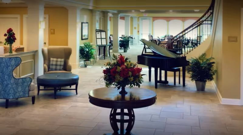 A bright interior lobby with a round table holding a flower arrangement, seating, a grand piano, and a curved staircase.