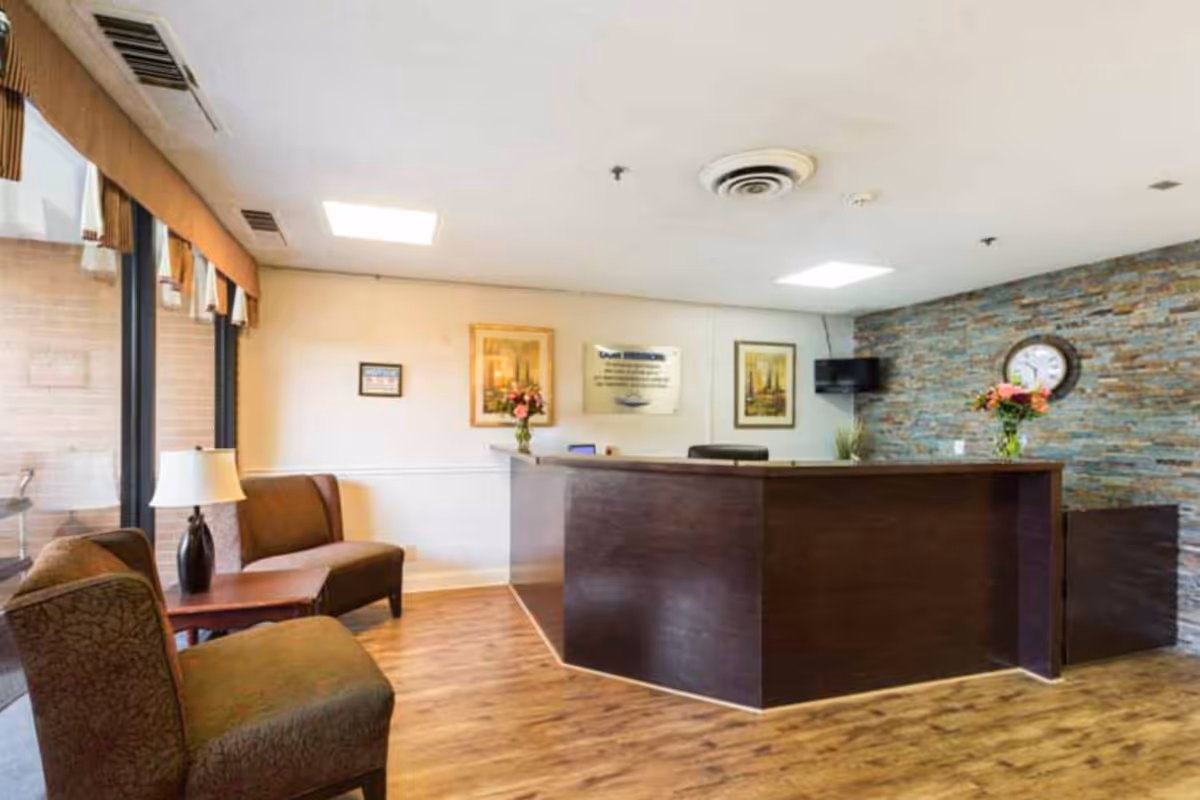 Reception area of Glenwood Health Center featuring a dark wood reception desk with flower arrangements, a wall clock, framed pictures, a small wall-mounted TV, and seating area with two upholstered chairs and a side table with a lamp near large windows.