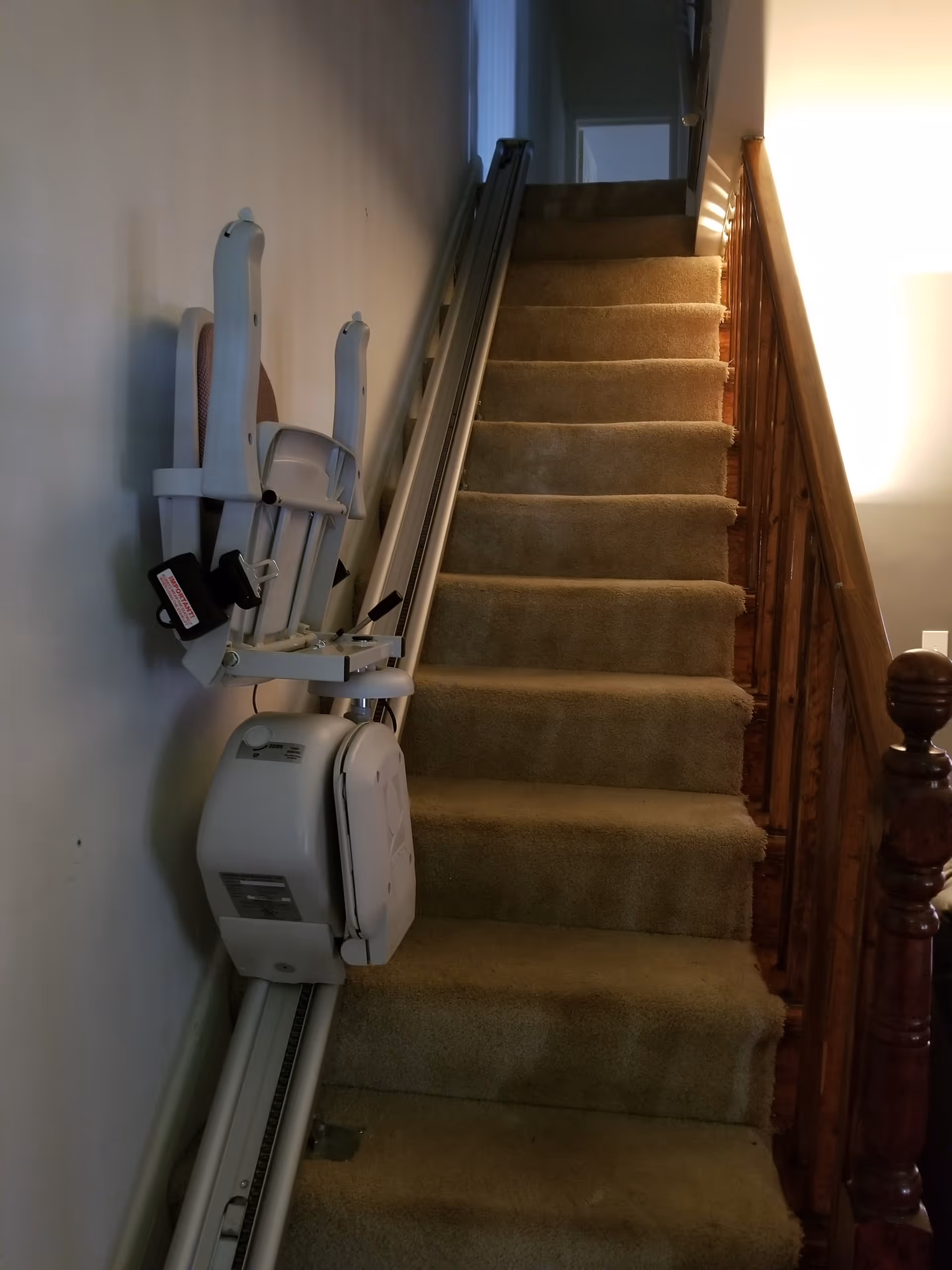 A carpeted staircase with a stairlift installed on the left side. The stairlift chair is folded up against the wall. The staircase has a wooden handrail on the right side and is lit by a lamp on the right wall.