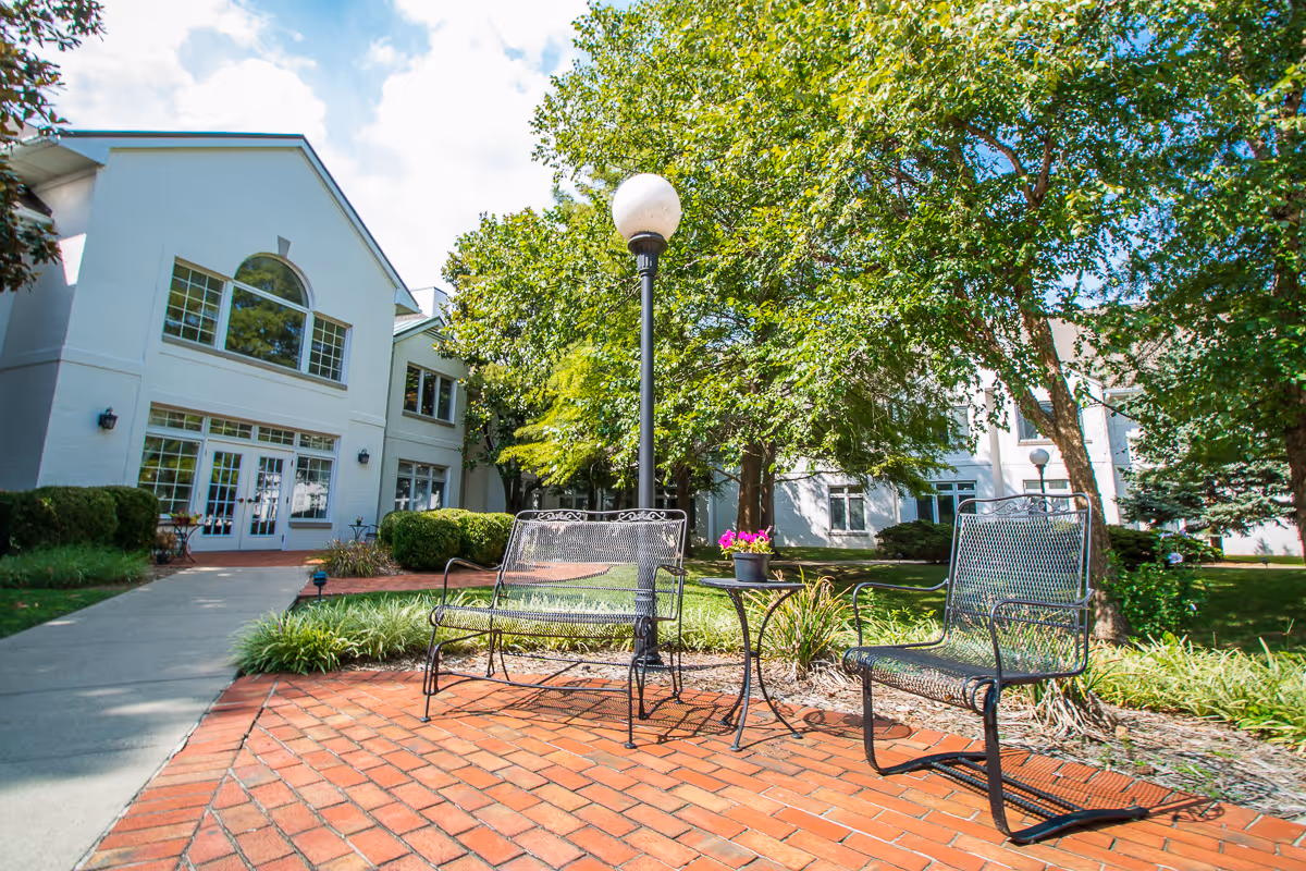 Outdoor seating area with two metal chairs and a small table with a potted plant on a brick patio. The area is surrounded by greenery and trees, with a white building in the background under a partly cloudy sky.