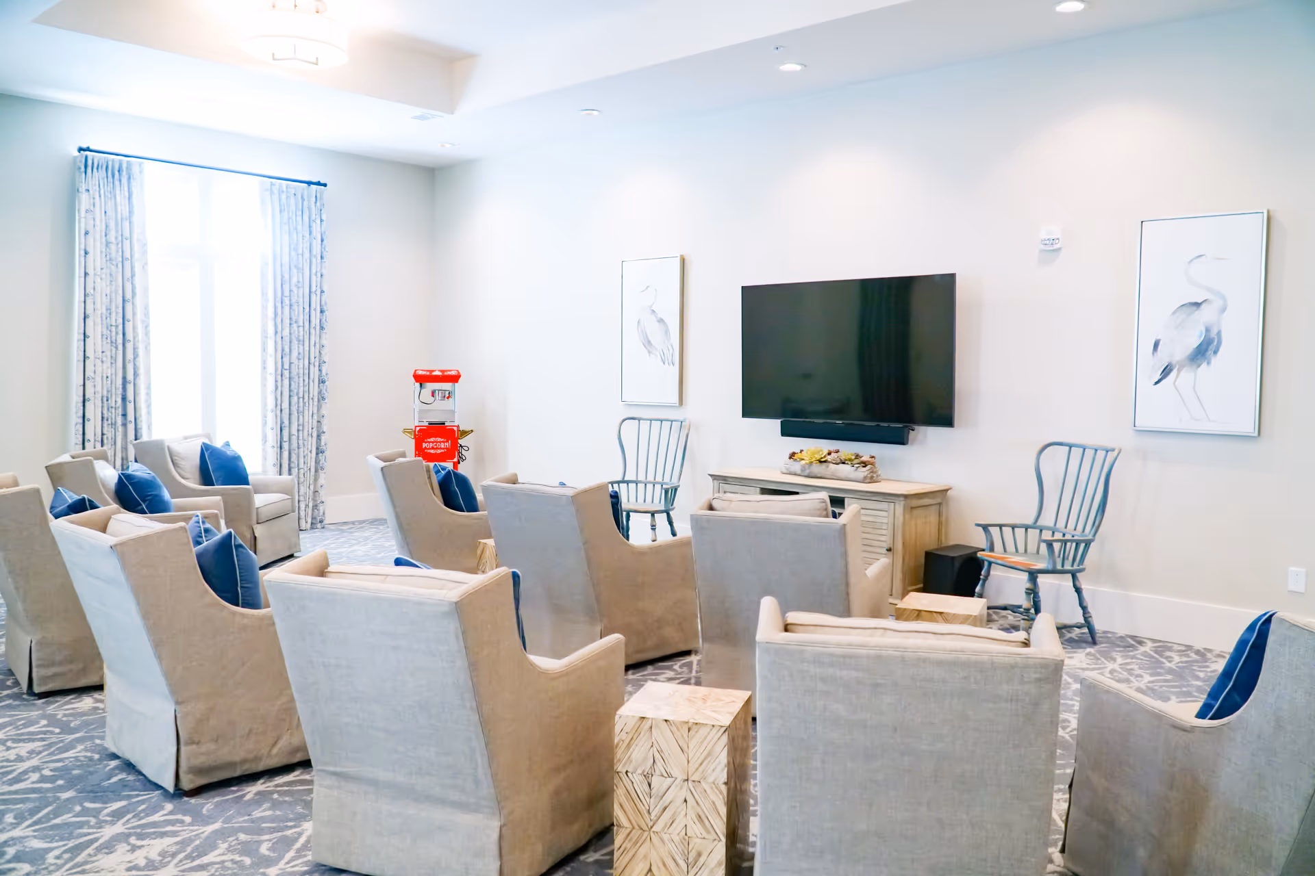 A bright and airy common living room area with multiple beige armchairs arranged in rows facing a wall-mounted flat screen TV. The room features patterned carpet, two framed bird artworks on the wall, a wooden TV console with decorative items, a red popcorn machine near a window with floral curtains, and two wooden chairs near the TV.
