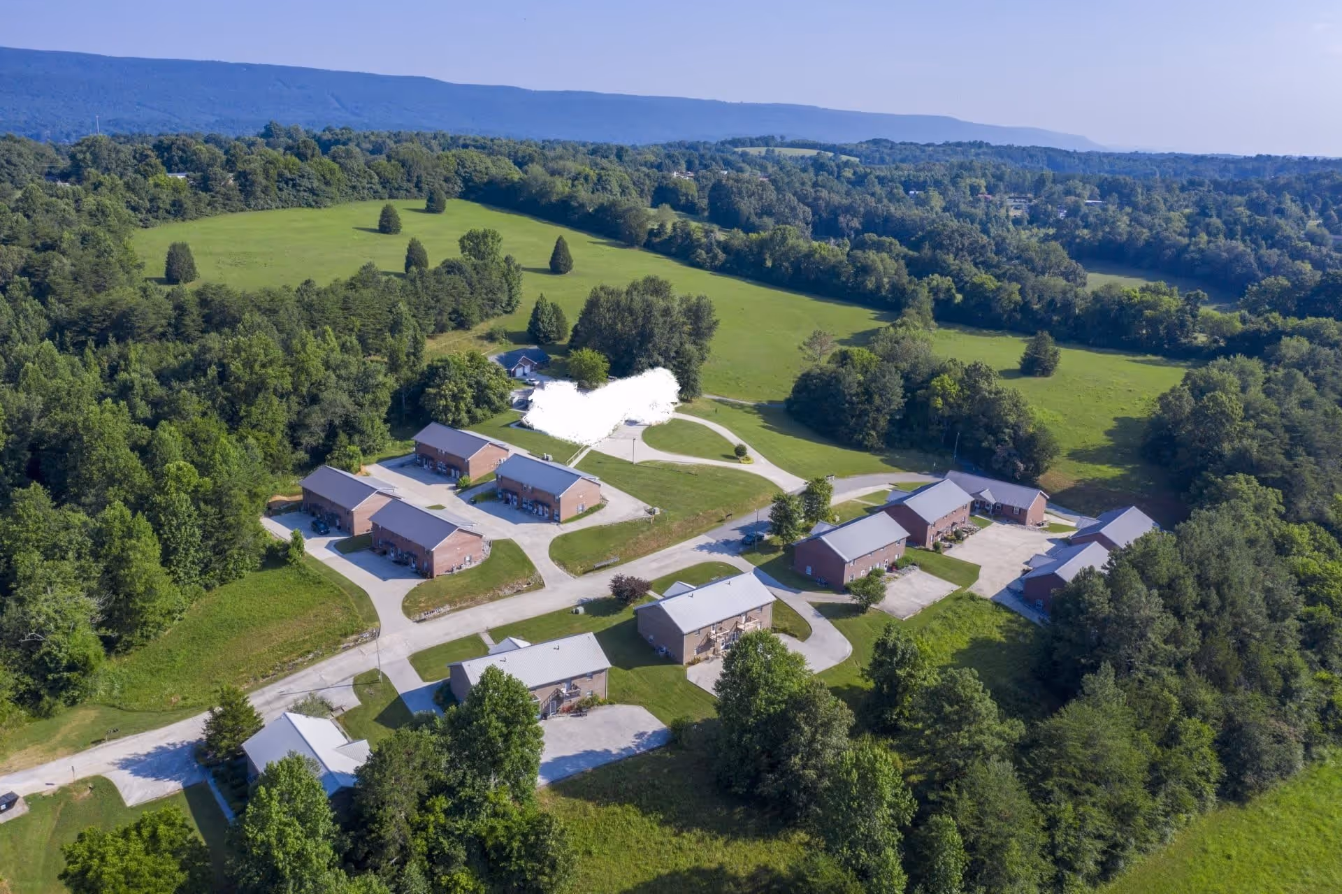 Aerial view of a senior living facility named Small Town Senior Living at Deer Ridge, showing multiple brick buildings with gray roofs arranged along paved roads surrounded by green lawns, trees, and open fields with hills in the background under a clear sky.