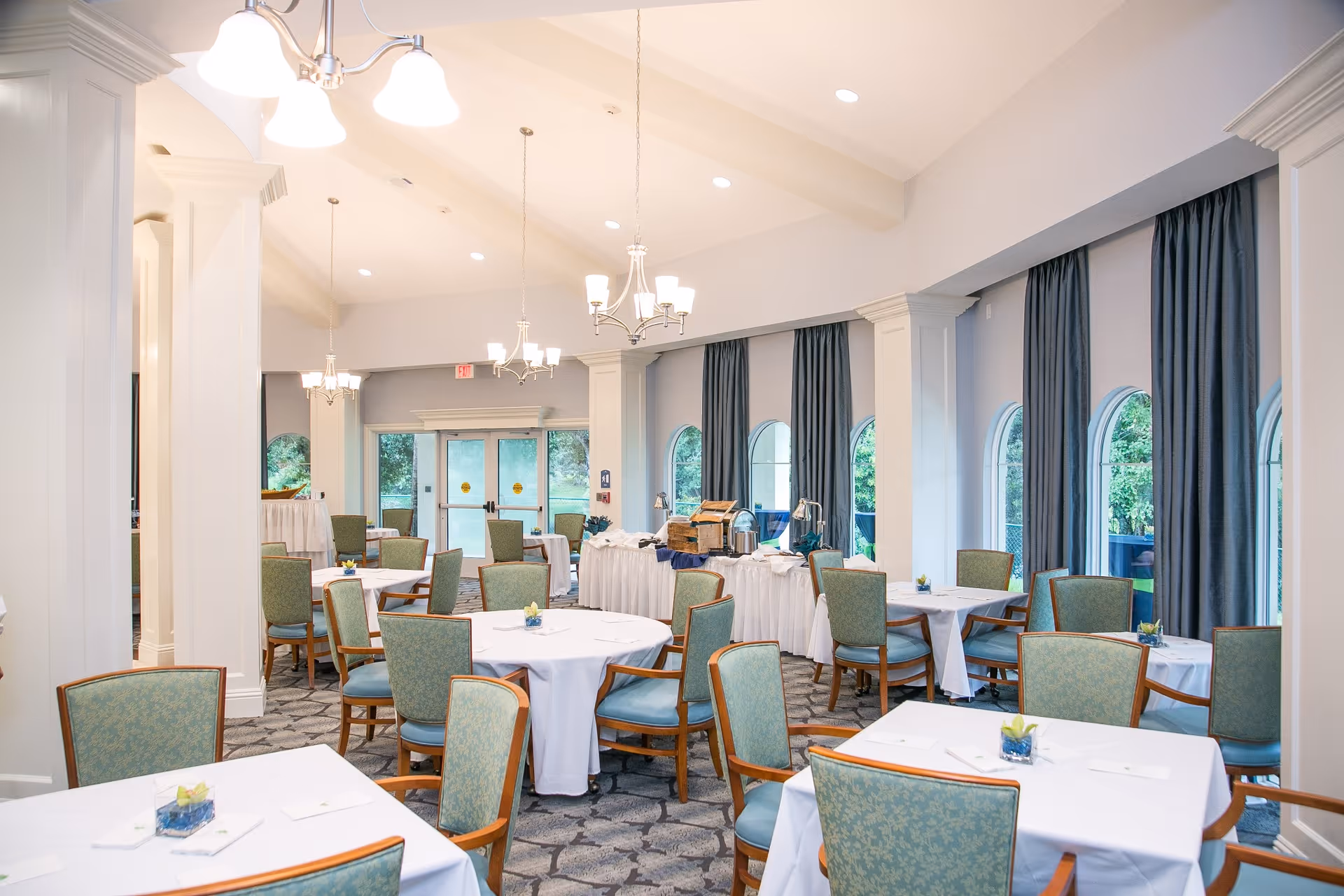 Bright dining room with round tables draped in white linens and green upholstered chairs under chandeliers and arched windows.