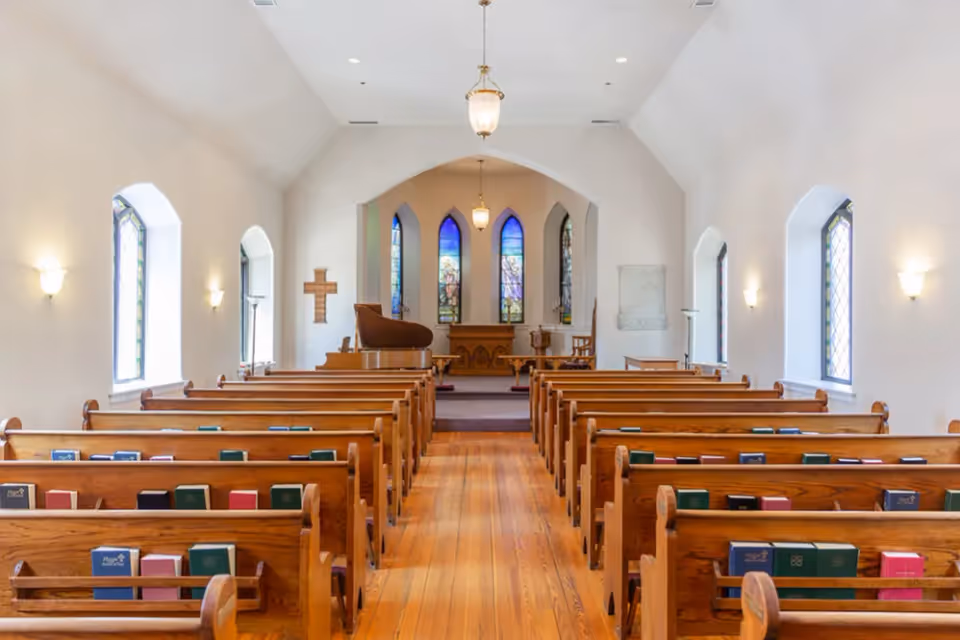 Interior view of a chapel with wooden pews arranged in rows facing an altar. The chapel features stained glass windows, a wooden floor, a grand piano, and a cross on the wall. Soft lighting fixtures are mounted on the white walls.