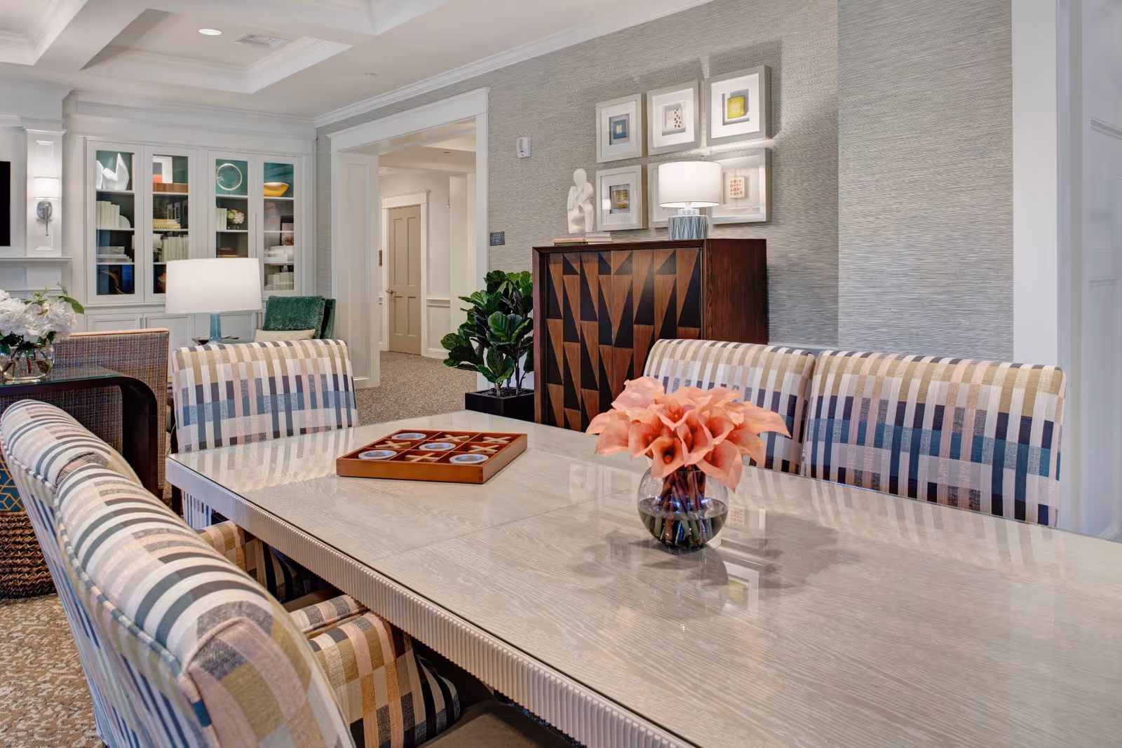 Bright communal dining area with a glossy table, patterned upholstered chairs, a vase of flowers, and decorative cabinets and artwork in the background.