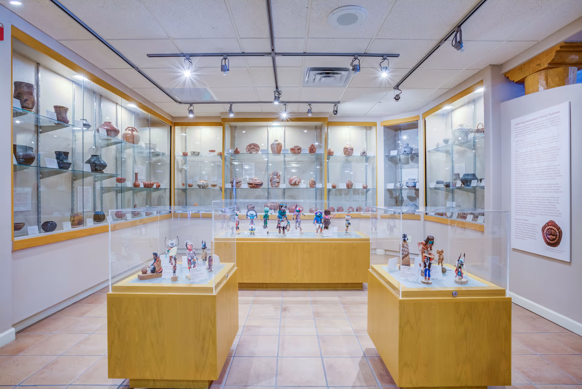 A well-lit museum-style room displaying various pottery and figurines in glass cases and on shelves along the walls. The room has tiled floors, a white ceiling with track lighting, and informational plaques on the walls.