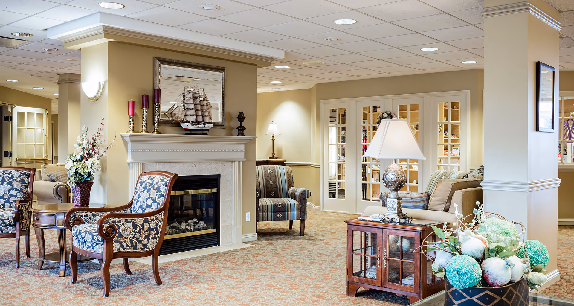 A cozy living room area in a senior living facility with patterned armchairs, a small wooden table, a fireplace with decorative candles and a model ship on the mantel, a lamp on a wooden cabinet, and floral arrangements. The room has beige walls, carpeted floor, and French doors leading to another room.