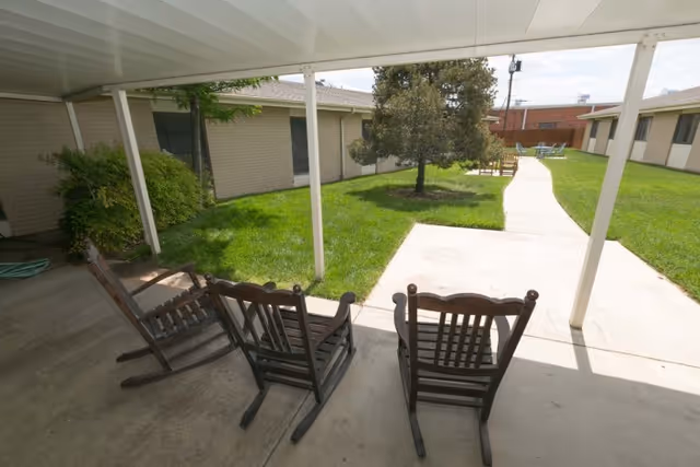 Three wooden rocking chairs under a covered patio overlooking a grassy courtyard with a walkway and surrounding single-story buildings.