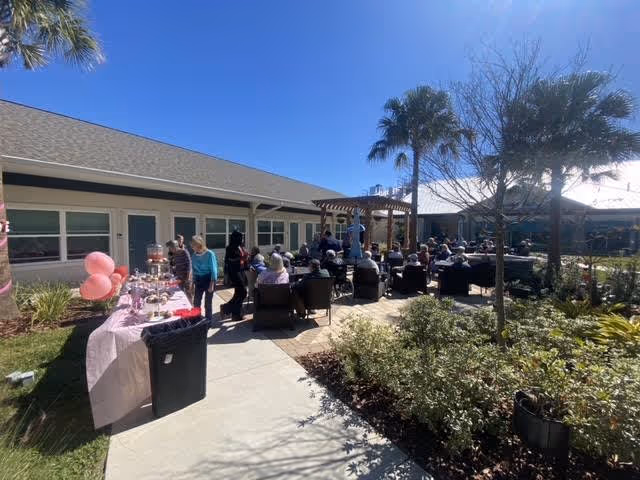 Residents and staff gathered in a sunny outdoor courtyard at the assisted living facility for a daytime social event with tables, chairs, palm trees, and pink balloons.