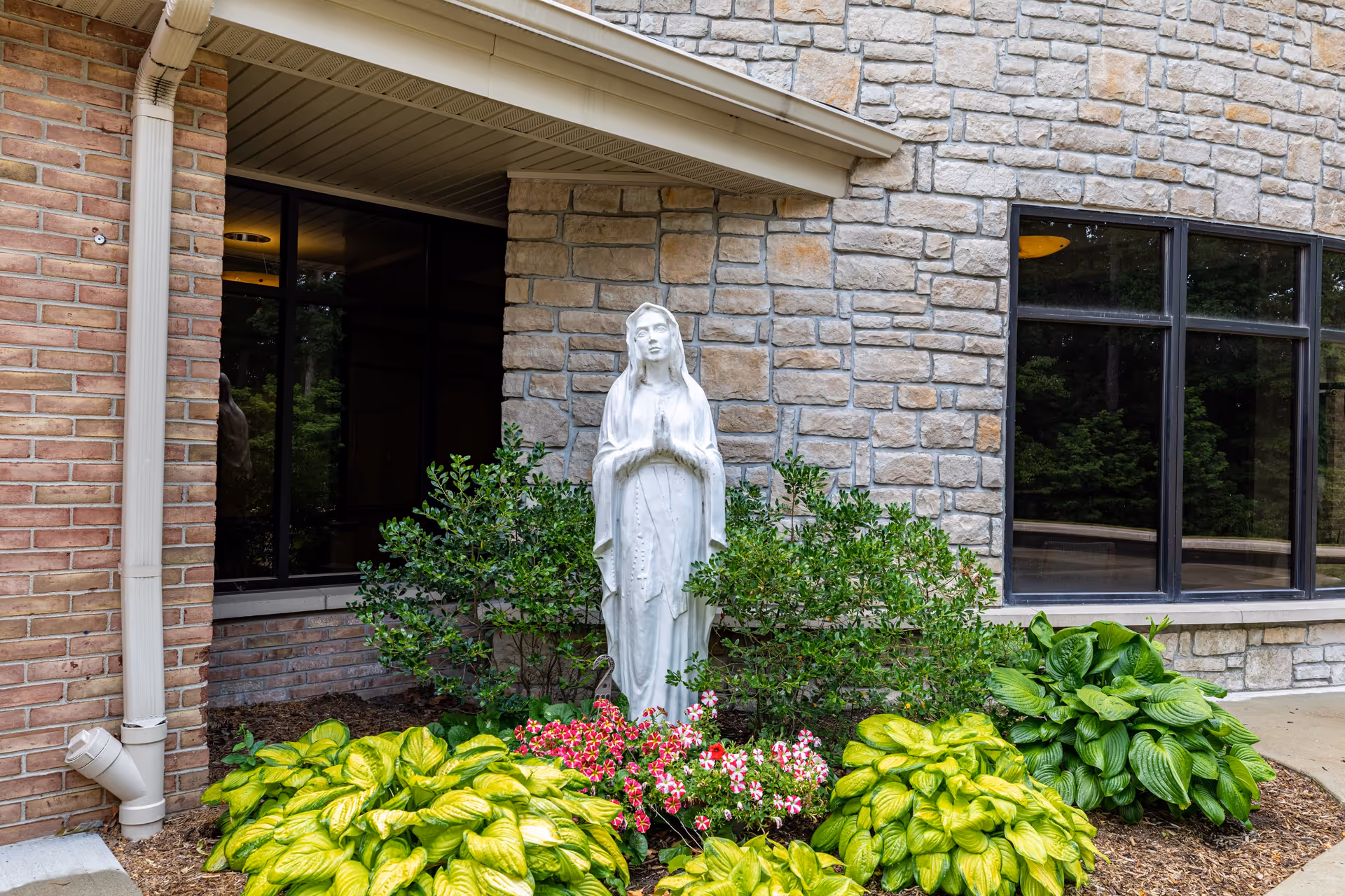 A white statue of a praying woman, likely the Virgin Mary, surrounded by green bushes and colorful flowers, positioned outside a stone and brick building with large windows.