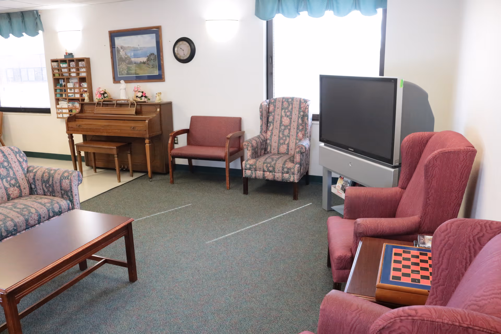 A cozy common sitting room with upholstered armchairs, a couch, coffee table, upright piano, and a television near a window.