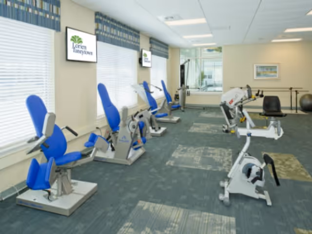 A fitness room with several exercise machines including stationary bikes and leg exercise equipment, featuring blue padded seats. The room has large windows with blinds and two screens on the wall displaying the Lorien Taneytown logo. The floor is carpeted and the walls are light-colored with a framed picture and a ballet barre in the background.