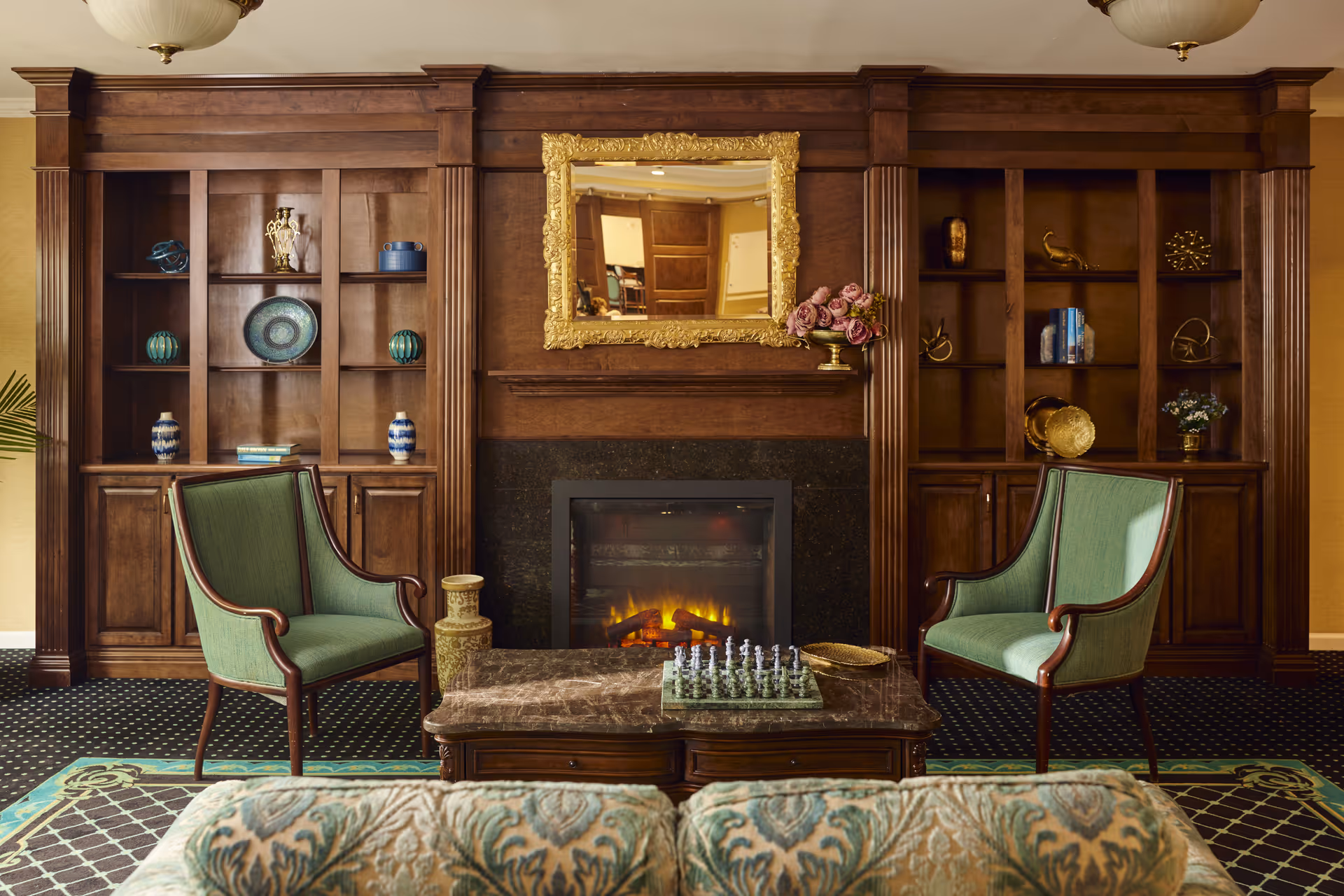 A cozy living room area with a dark wooden fireplace mantel and built-in shelves on either side. The fireplace has a glowing fire and above it hangs an ornate gold-framed mirror. Two green upholstered armchairs face a marble coffee table with a chess set on it. The room features patterned carpet and a floral upholstered sofa partially visible in the foreground.