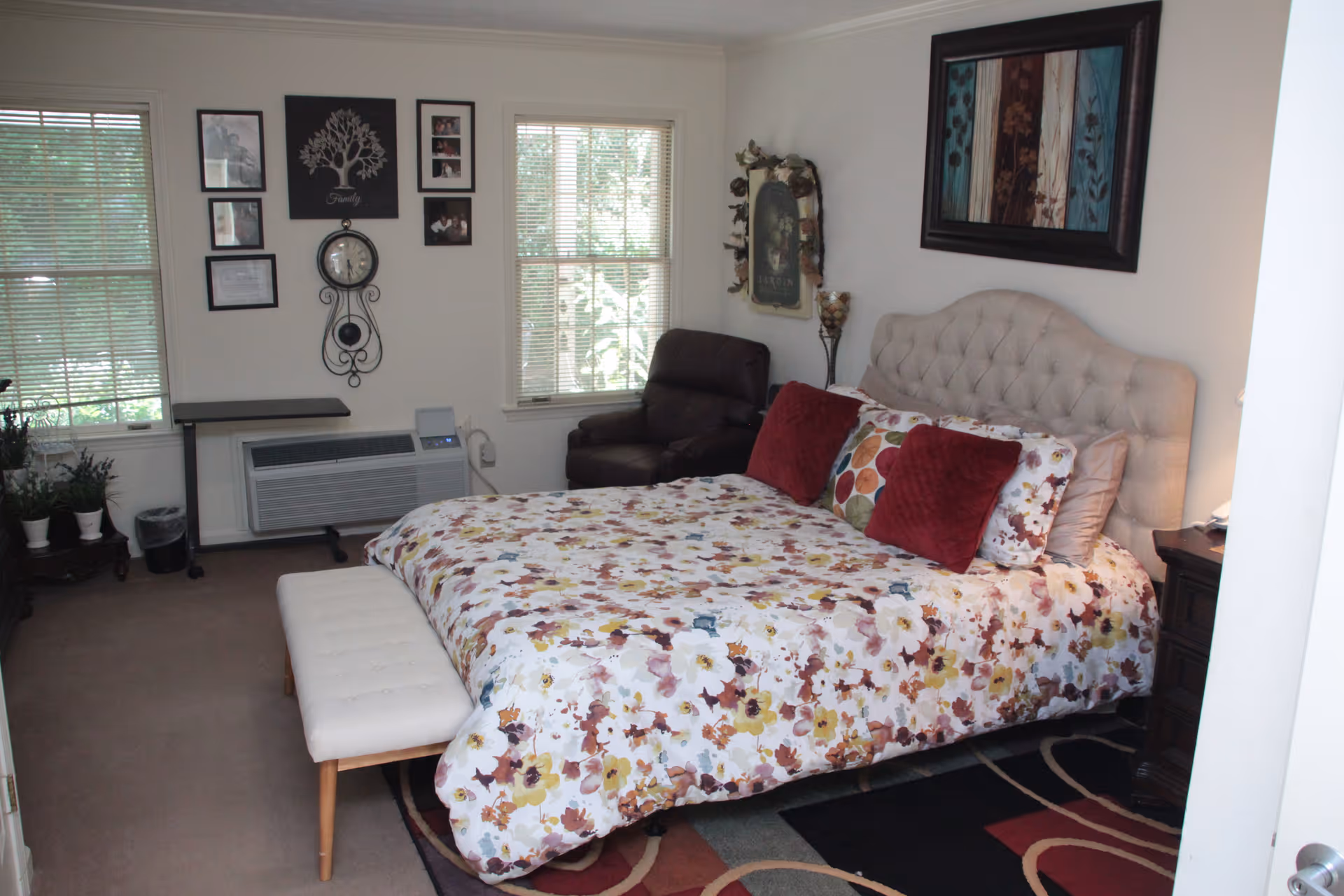 Bright bedroom featuring a bed with a floral comforter and tufted headboard, a bench at the foot, an armchair, windows, and wall decor.