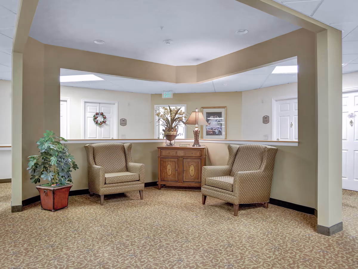 A cozy seating area in a senior living facility with two patterned armchairs facing each other, a wooden cabinet with a lamp and a plant on top, and a potted plant on the floor. The walls are beige, and there are white doors and framed artwork in the background.