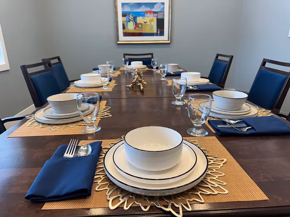 A long dining table set for eight with white plates, bowls, glasses, silverware wrapped in blue napkins, and decorative placemats. The room has gray walls, a window on the left, and a colorful framed painting on the far wall.