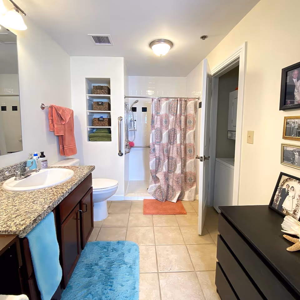 Well-lit bathroom with a granite vanity and sink, toilet, built-in shelves, a walk-in shower with a patterned curtain, and a laundry closet.