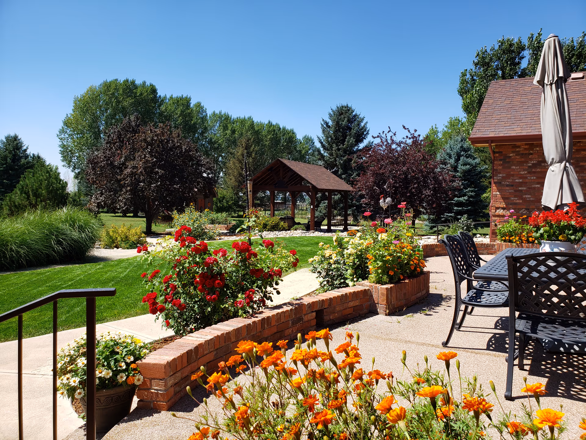 Sunny outdoor patio and garden with colorful flowers, a gazebo, and metal patio seating beside a brick building.