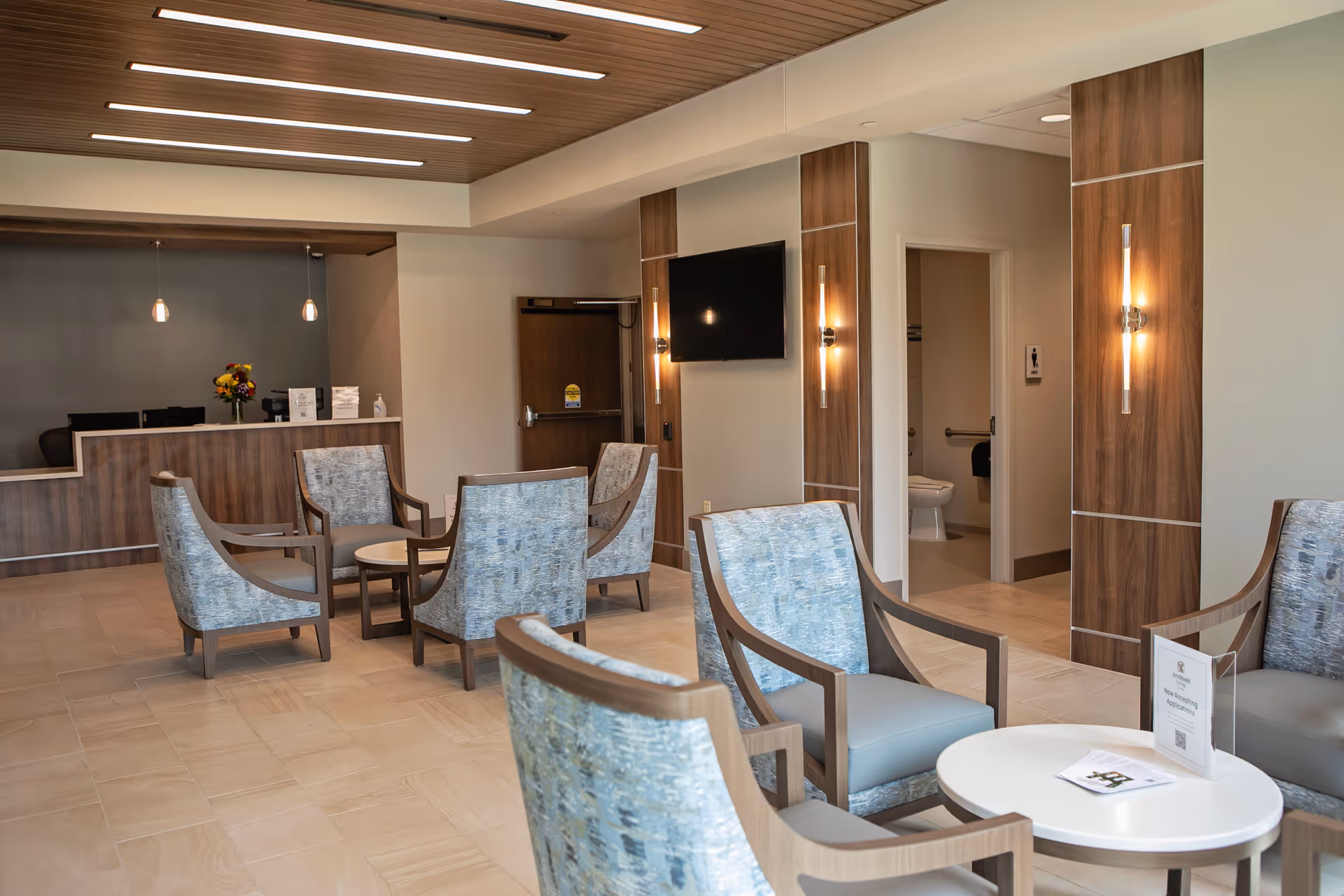 A modern reception lobby with upholstered chairs around small tables, a front desk, and a wall-mounted TV.