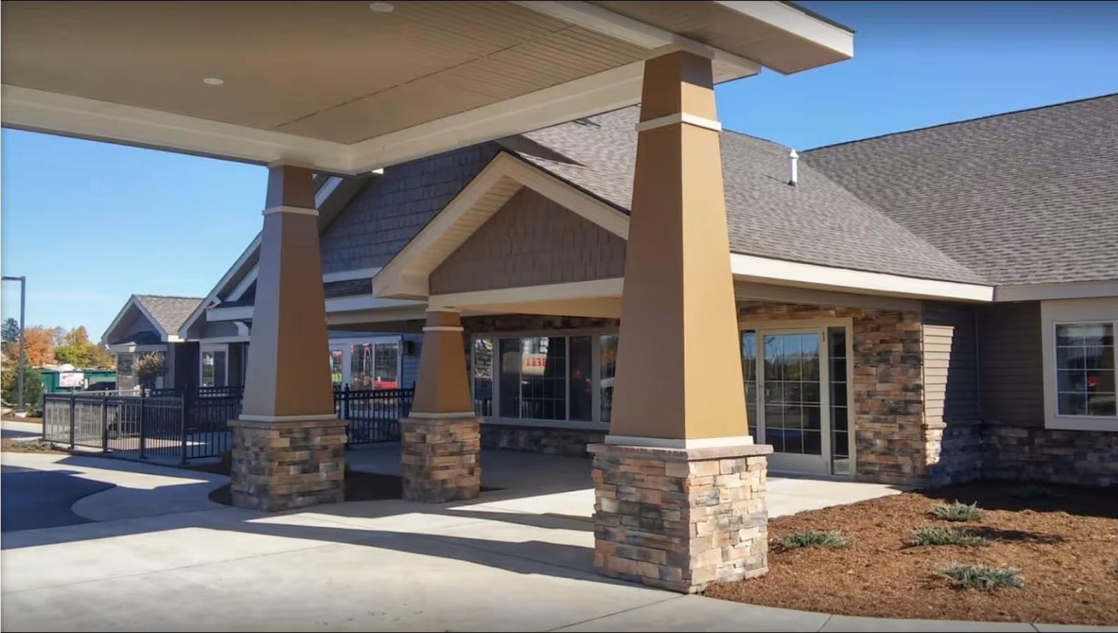 Covered entrance area of a senior living facility with stone and beige pillars, a sloped roof, and large windows and doors. There is a paved driveway and some landscaping with small plants and mulch.