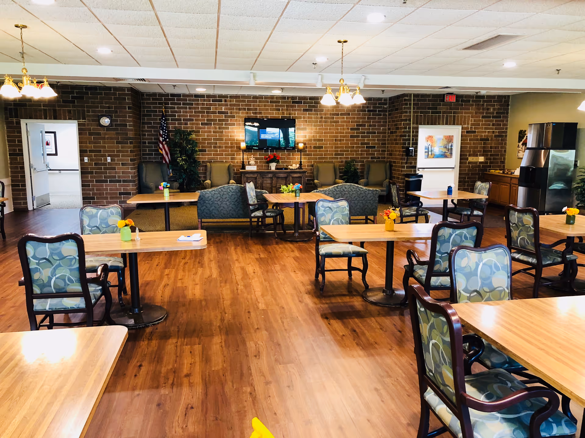 Spacious assisted living dining/common area with wooden tables and patterned chairs facing a brick accent wall and TV.