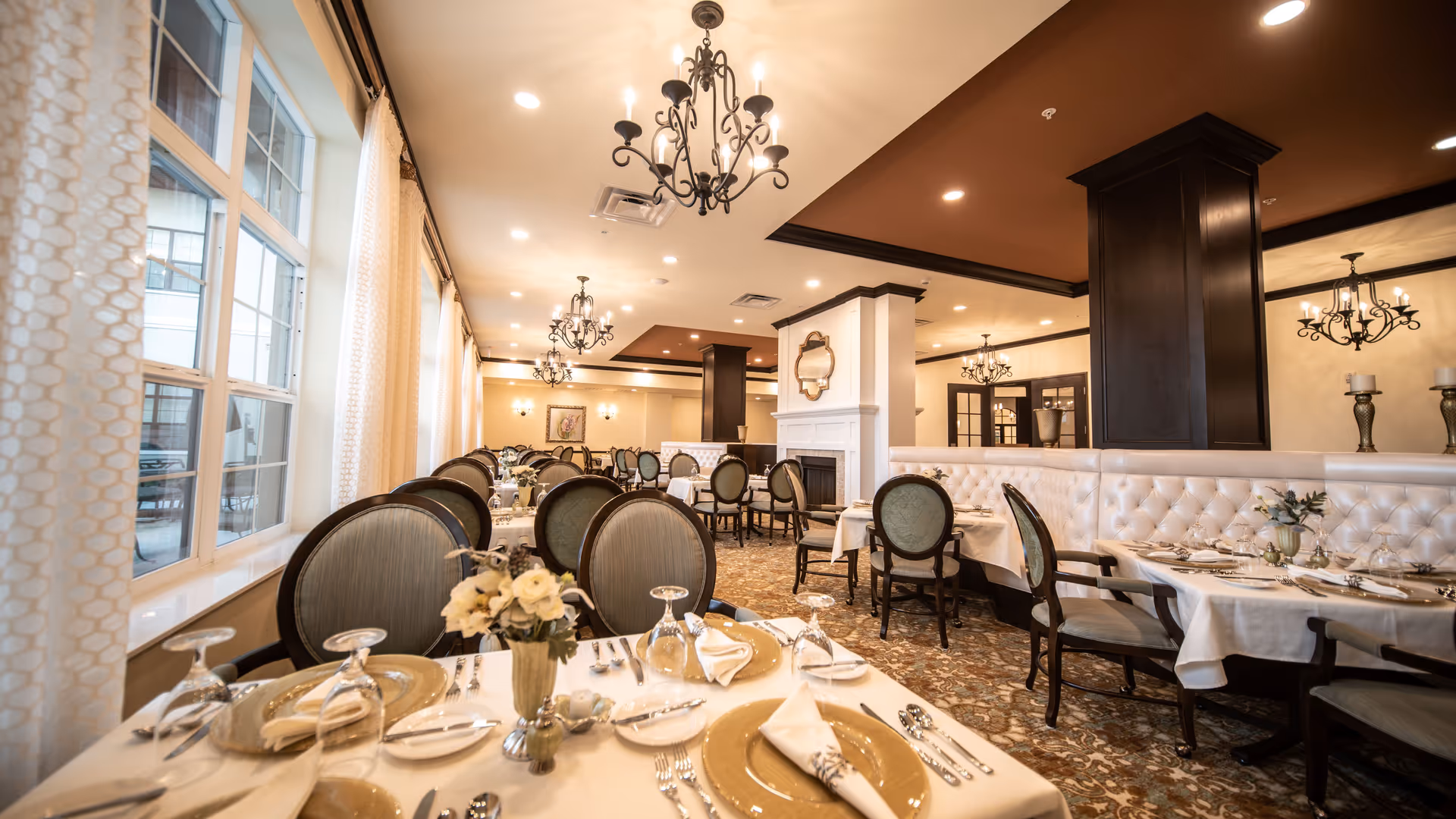 Elegant dining room with multiple tables set with gold charger plates, white napkins, silverware, and glassware. The room features upholstered chairs, chandeliers, large windows with curtains, and a fireplace with a mirror above it. The carpet has a patterned design and the ceiling has recessed lighting and dark wood accents.