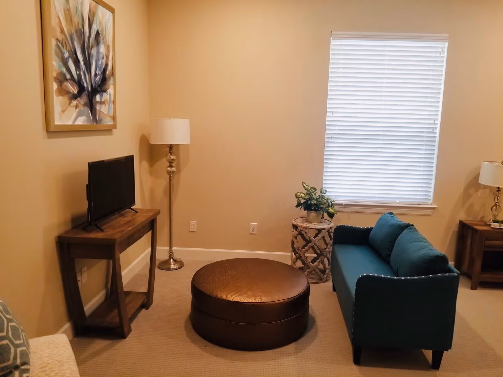 A cozy living room with beige walls and carpet. The room features a teal upholstered sofa with two cushions, a round brown ottoman in the center, a wooden TV stand with a flat-screen TV, a tall floor lamp with a white shade, a small decorative side table with a potted plant, and a window with white blinds. A framed abstract artwork hangs on the wall above the TV stand.