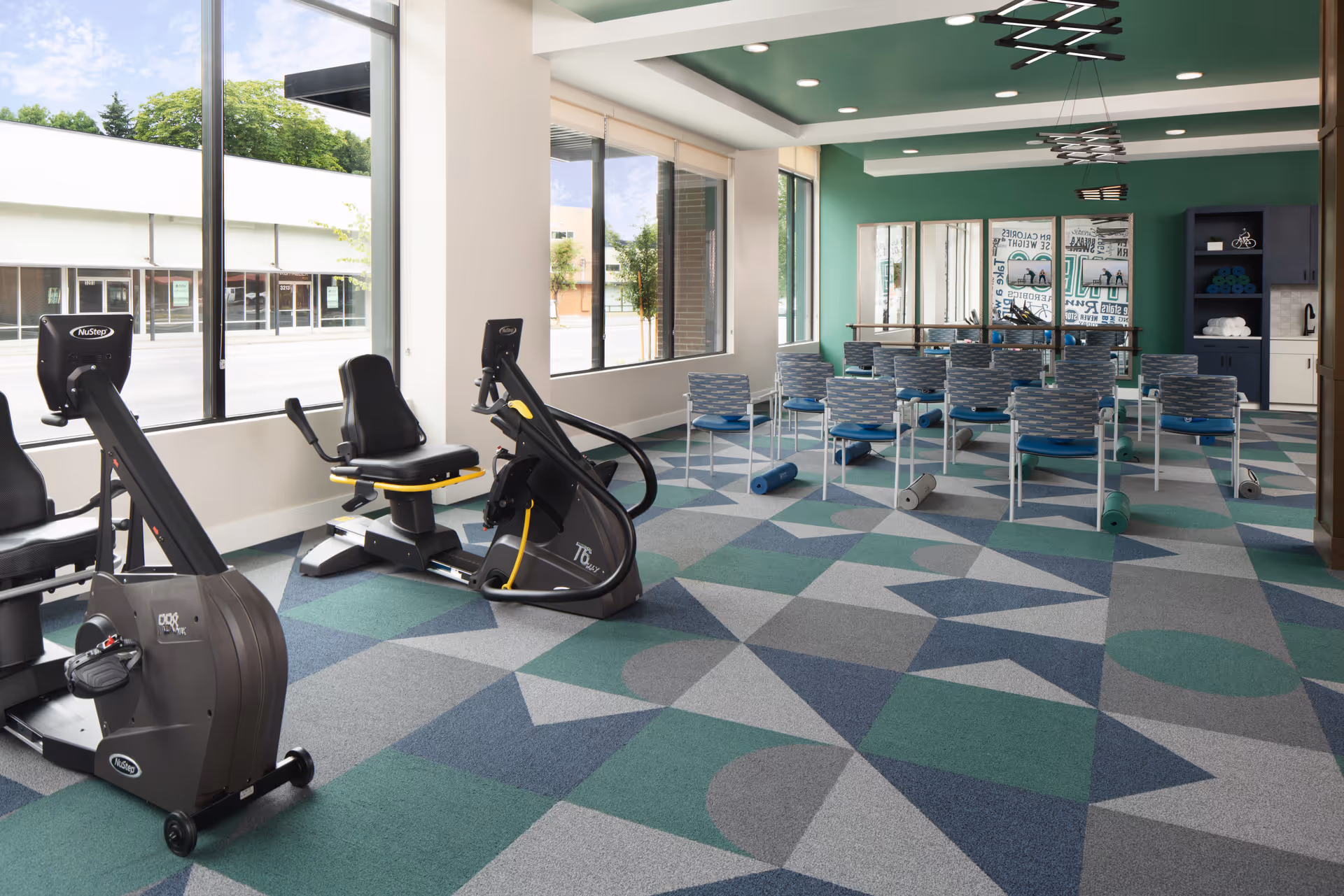 A bright exercise room with large windows letting in natural light. The room features two stationary exercise bikes on the left side and a row of chairs arranged in front of mirrors on the far wall. Several yoga mats are rolled up and placed near the chairs. The floor has a geometric patterned carpet in shades of green, blue, and gray. The walls are painted green and white, and there are modern light fixtures on the ceiling.