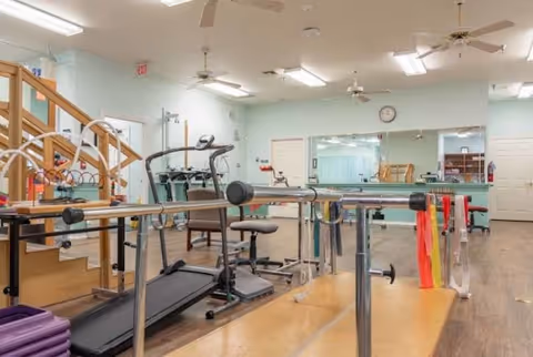 Interior view of a fitness and rehabilitation room in an assisted living facility featuring exercise equipment such as a treadmill, parallel bars for walking support, resistance bands, and chairs. The room has light-colored walls, ceiling fans, fluorescent lighting, a large mirror on one wall, and wooden flooring.
