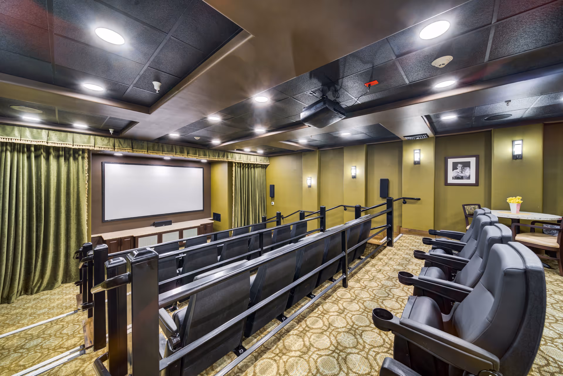 Interior view of a small theater room with two rows of black cushioned seats with armrests and cup holders facing a large blank projection screen. The room has green walls, patterned carpet, recessed ceiling lights, and a ceiling-mounted projector. There is a small table with chairs and a popcorn container in the back corner.